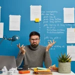stressful-professional-male-geek-concentrated-monitor-modern-laptop-wears-optical-glasses-poses-coworking-space-against-blue-background